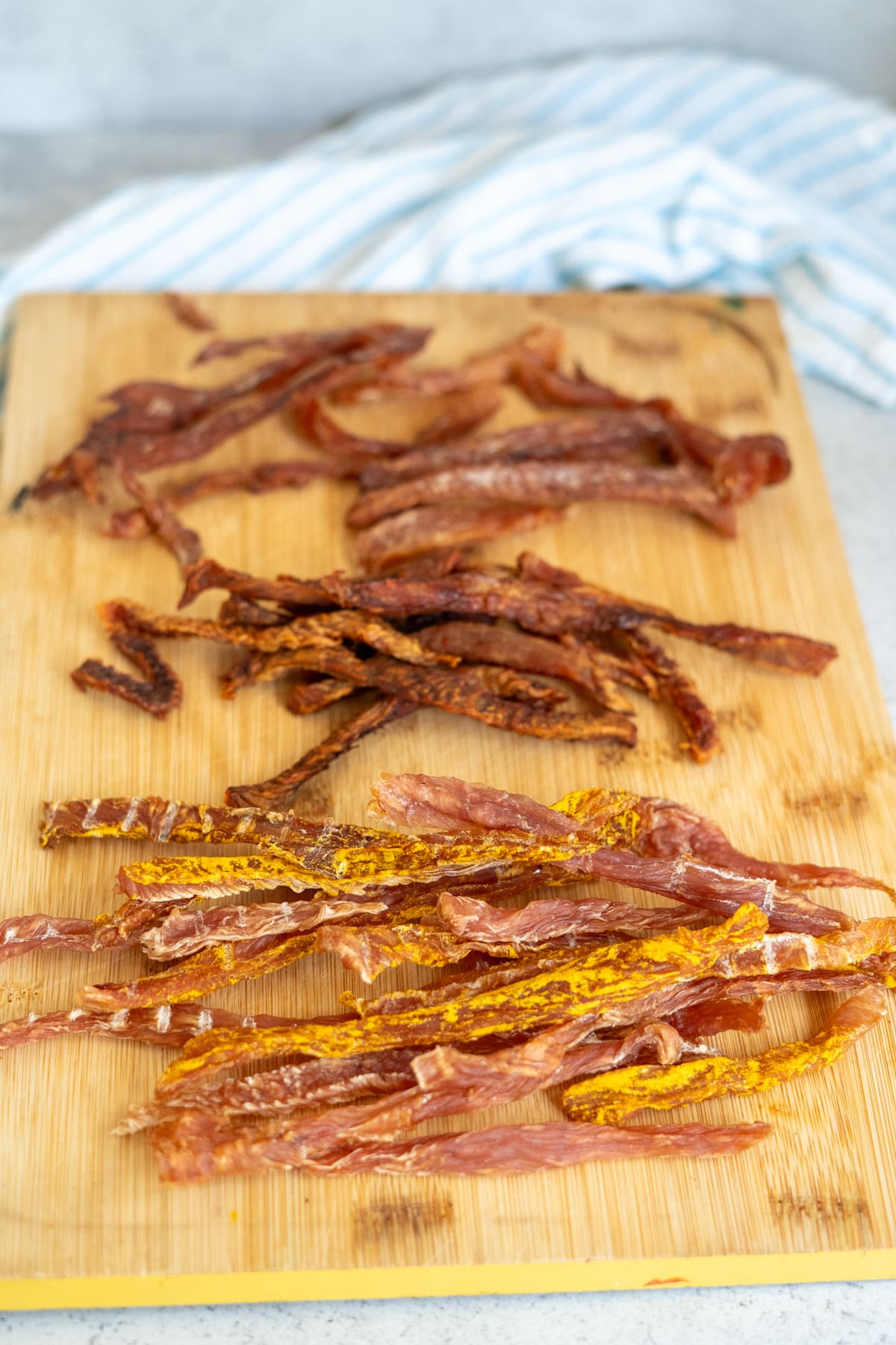 Three varieties of dried meat strips, including turkey jerky for dogs, are arranged on a wooden chopping board, with a striped cloth in the background.