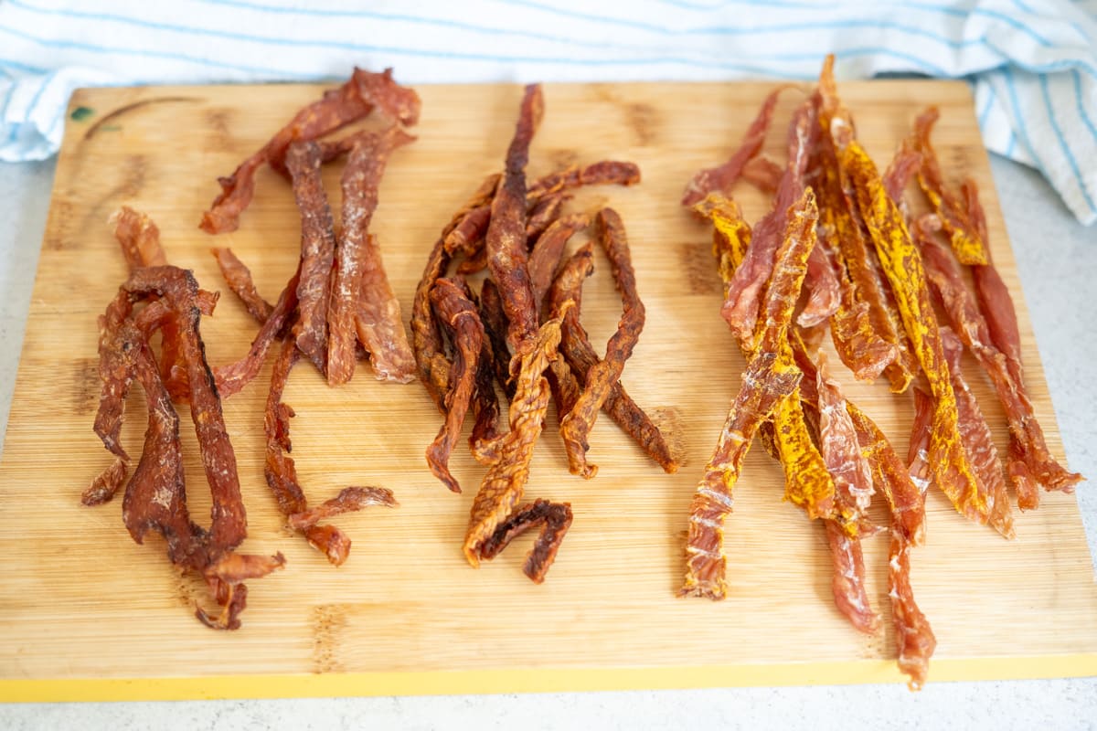 Three groups of dried meat strips, including turkey jerky for dogs, are arranged on a wooden chopping board, with a blue and white striped cloth in the background.