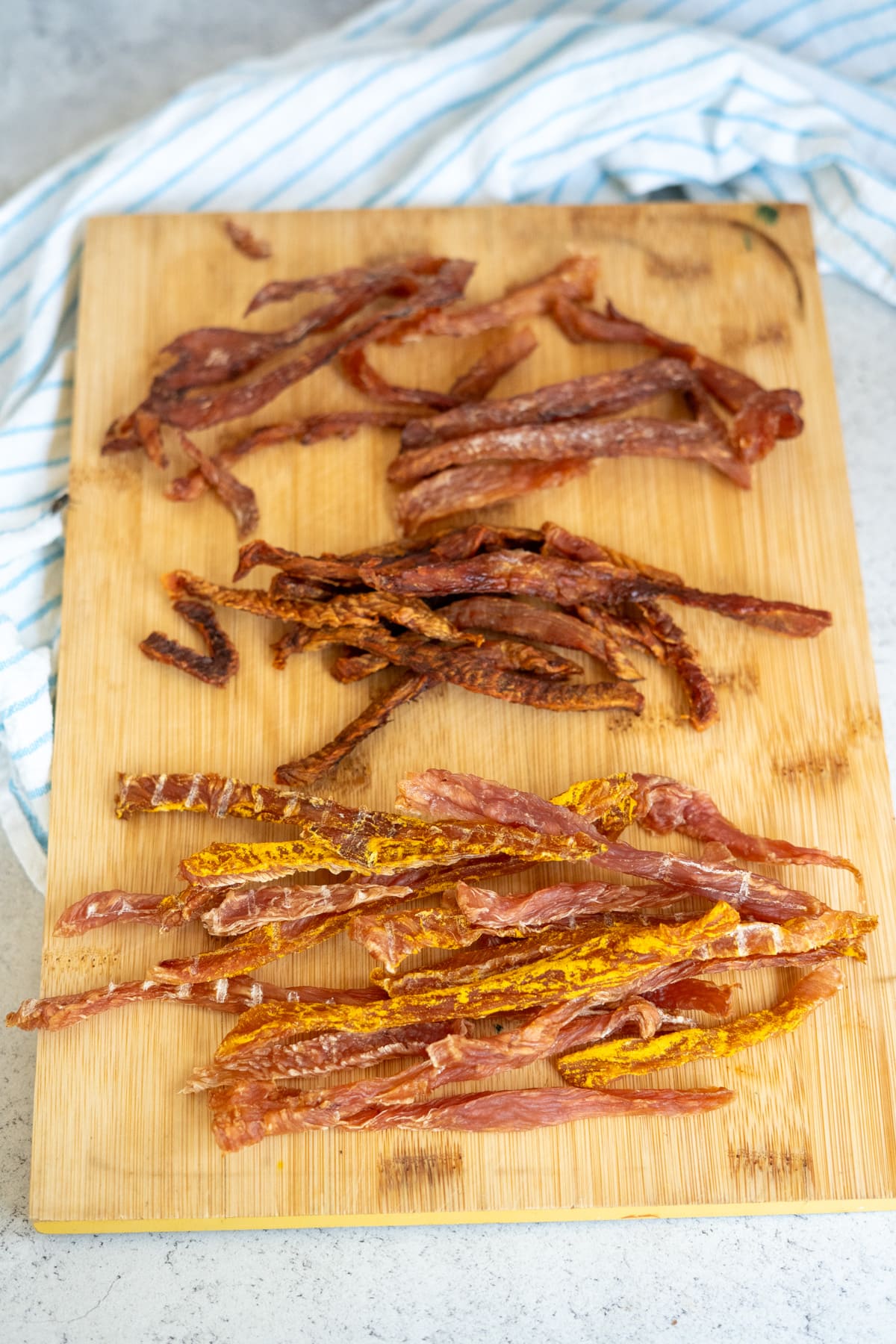 Three piles of sliced, dried jerky—perfectly suited as turkey jerky for dogs—are arranged on a wooden chopping board, with a striped cloth partially visible in the background.