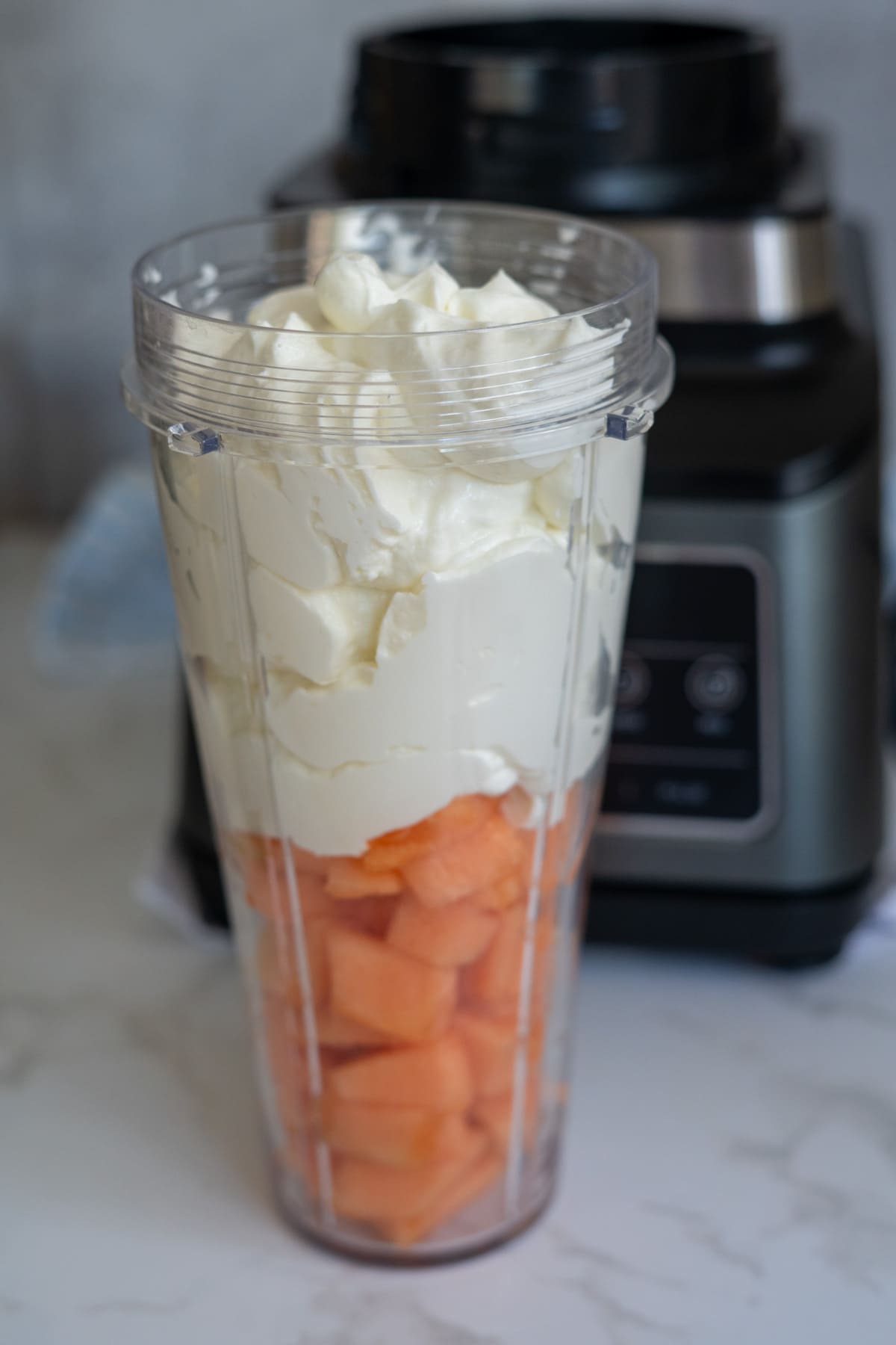 A blender jug filled with cubed cantaloupe and topped with a large amount of yoghurt, with a blender base in the background.