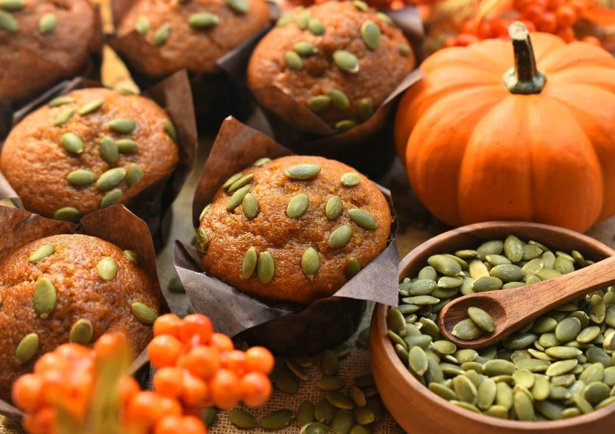 Pumpkin muffins topped with pumpkin seeds sit next to a small pumpkin, orange berries, and a bowl of pumpkin seeds with a wooden spoon.