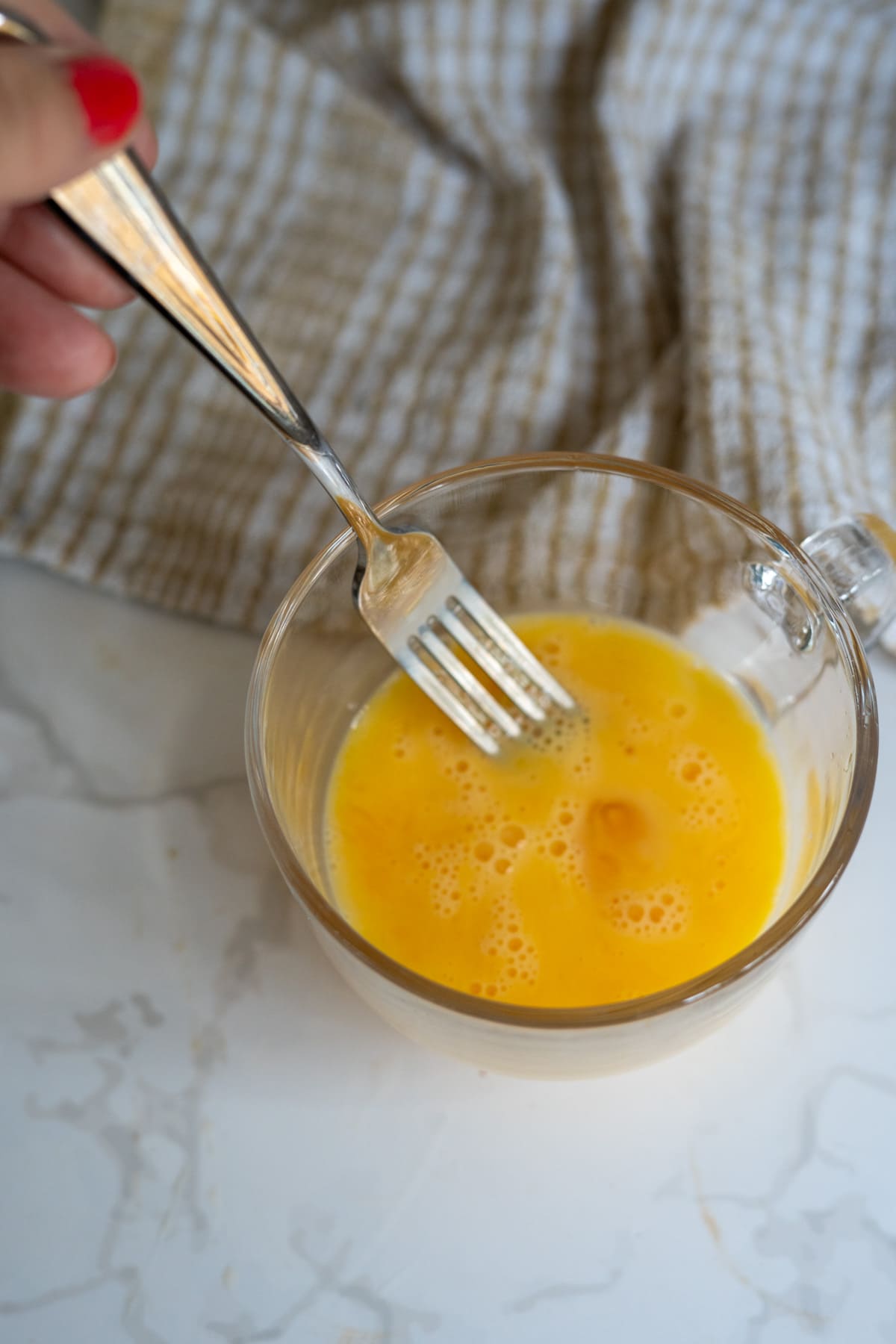 A hand uses a fork to whisk scrambled eggs for dogs in a clear glass bowl on a marble worktop, with a beige tea towel in the background.