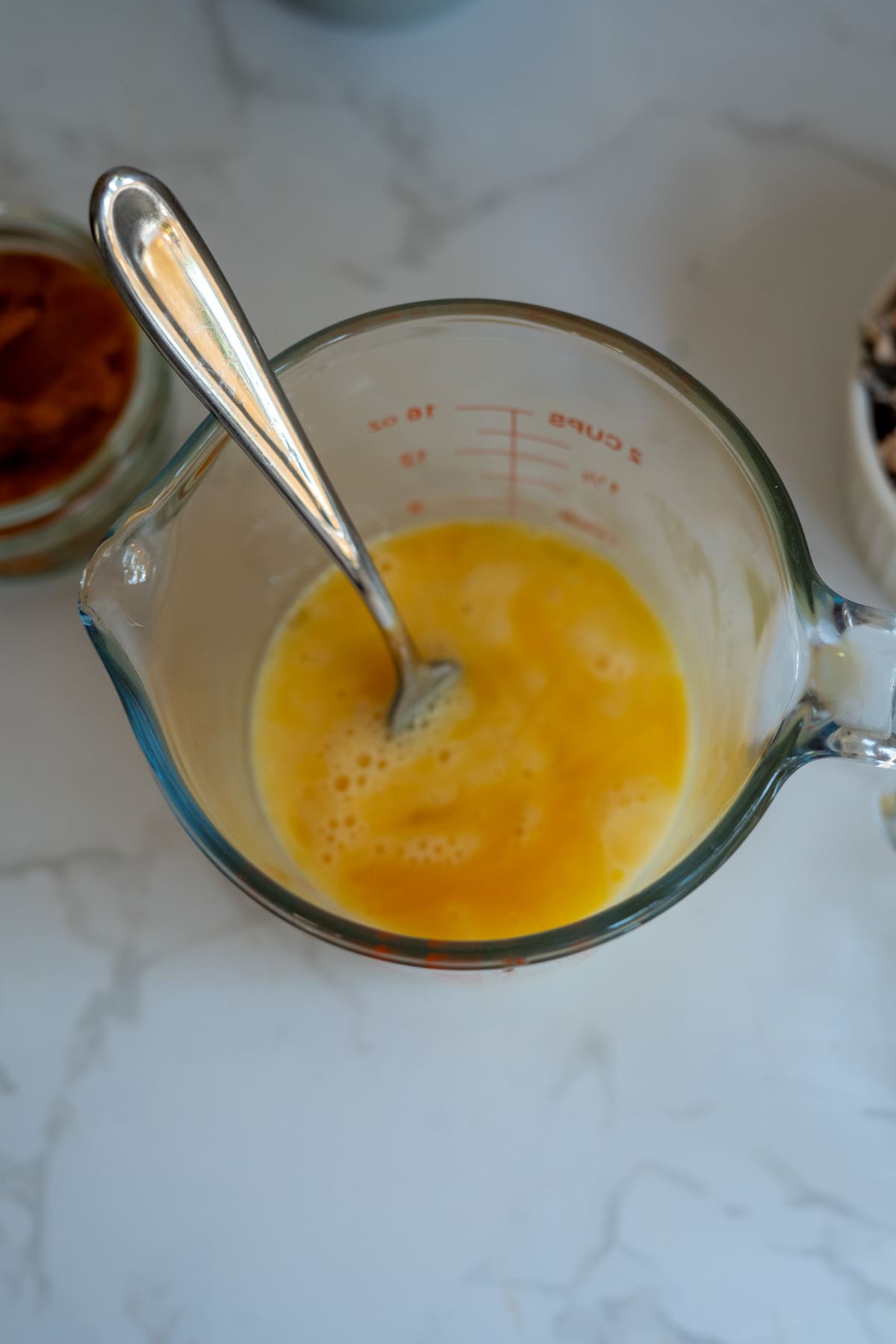 A glass measuring jug with beaten eggs and a fork inside is on a white worktop.