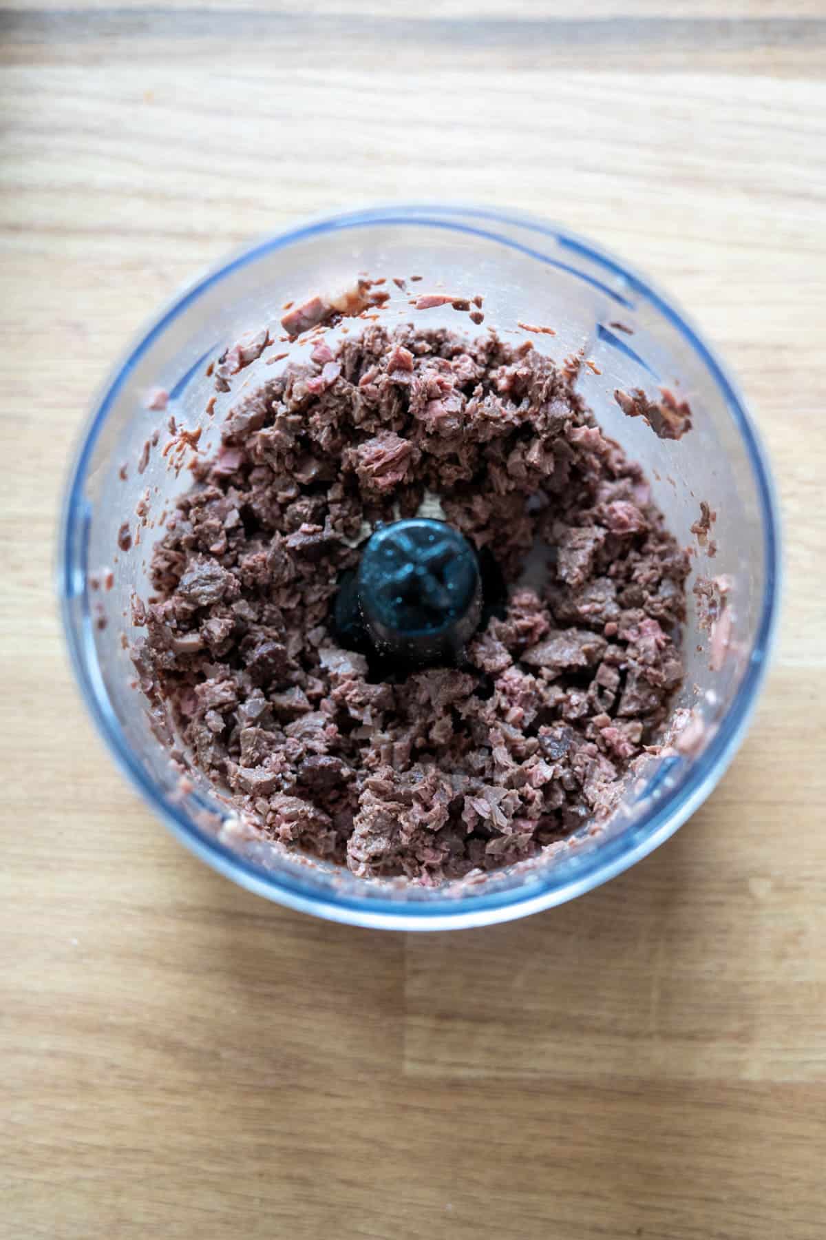 Chopped black olives in a food processor bowl on a wooden surface.