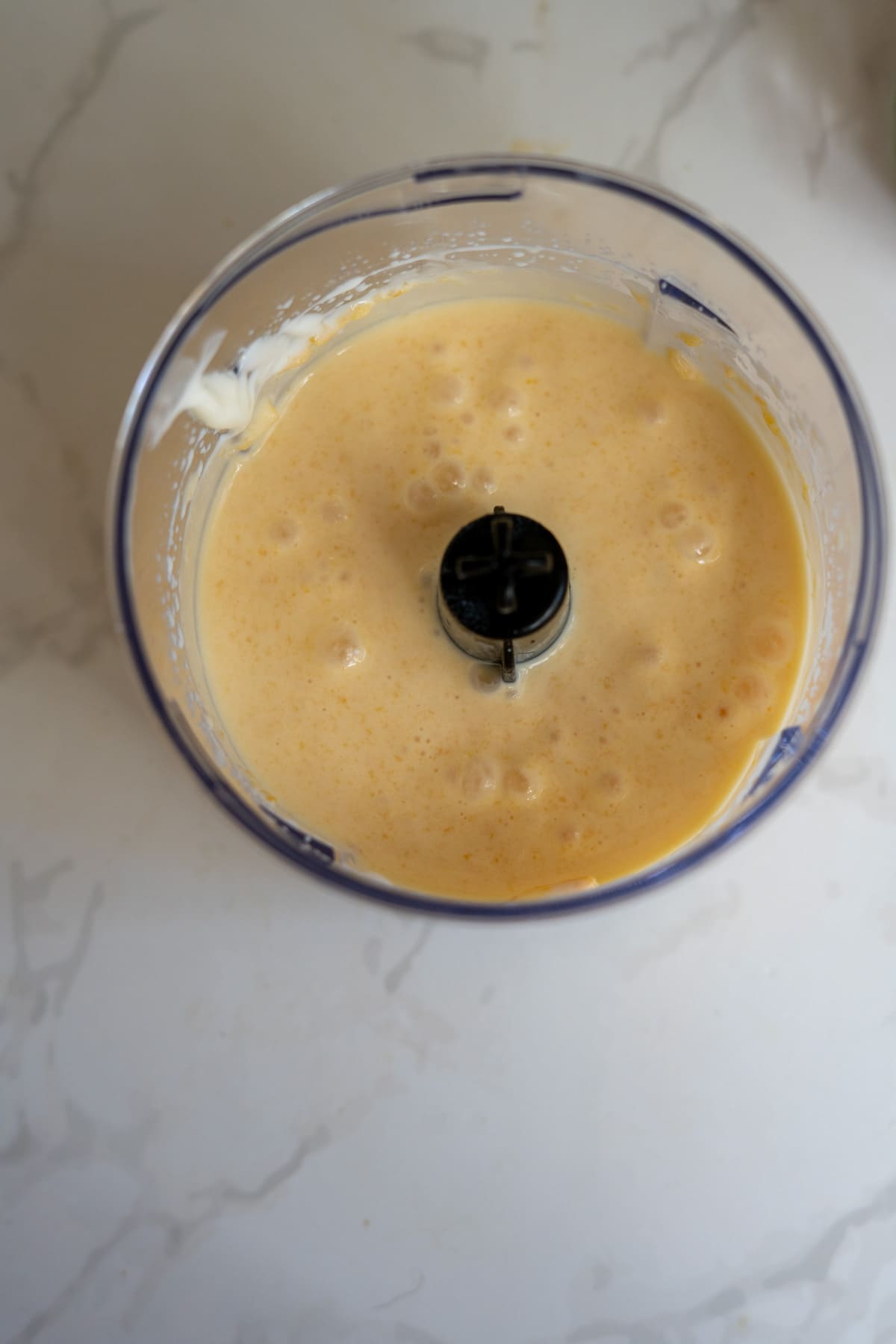 Blended yellow mixture with bubbles inside a transparent food processor bowl on a white marble surface, perfect for making Frozen Peach Raspberry Dog Treats.