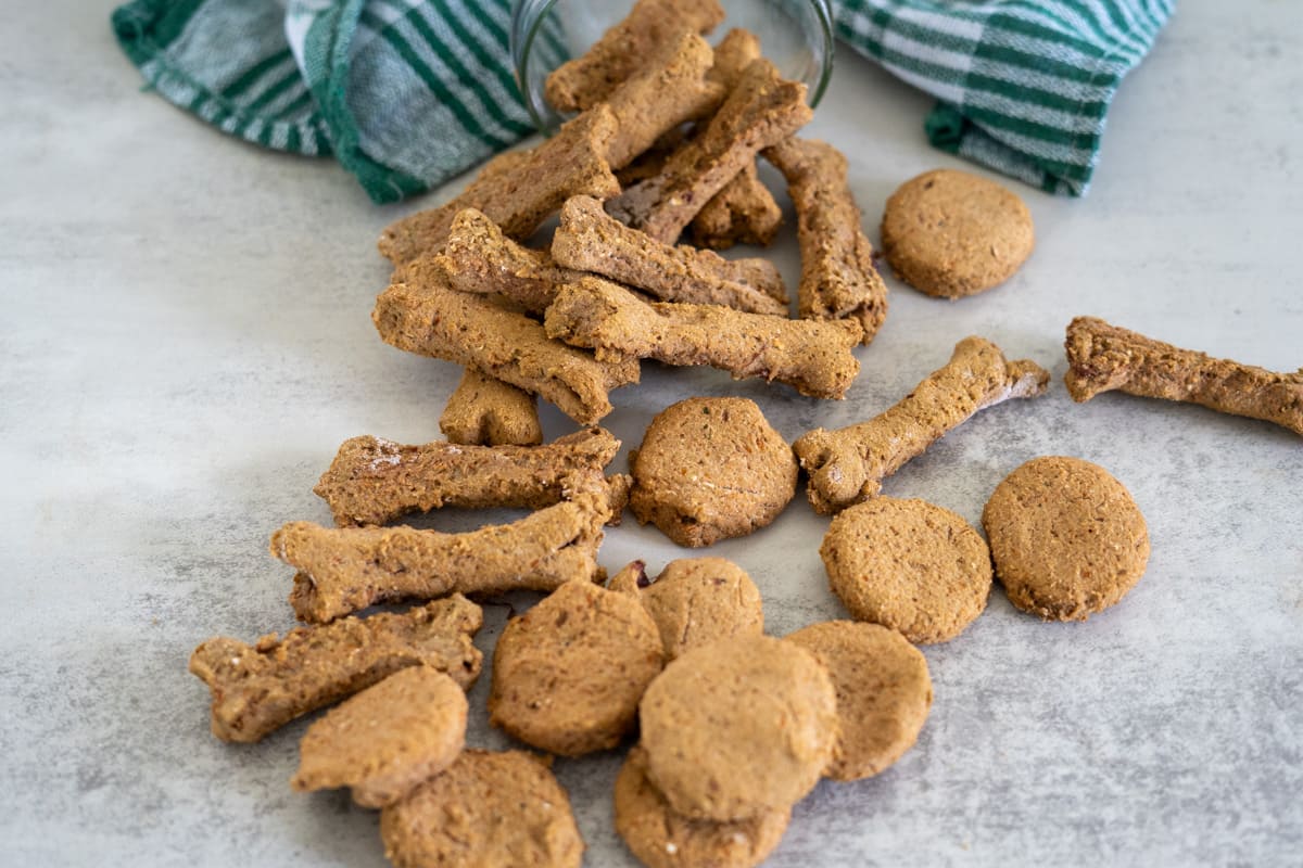 Homemade liver dog treats shaped like bones and circles are scattered on a light surface near two green and white striped towels.