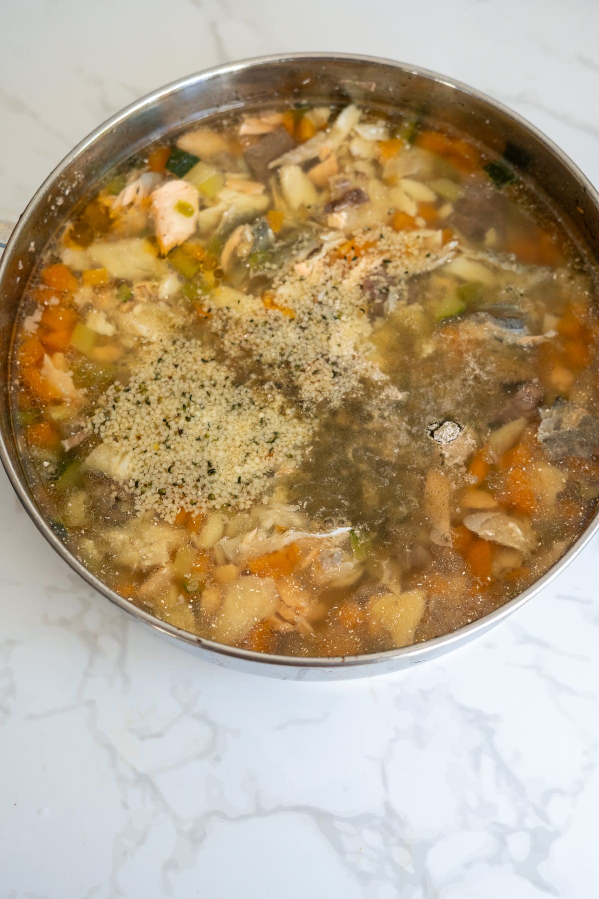 A large metal saucepan filled with chopped vegetables, chicken pieces, and seasonings in clear stock—perfect for a nourishing meal or as inspiration for a wholesome fish stew for dogs—sitting on a marble worktop.