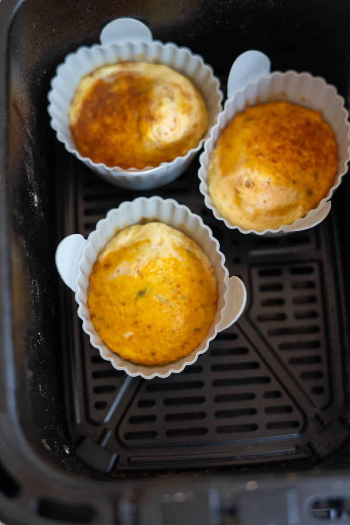 Three browned egg muffins in white silicone moulds inside an air fryer basket.