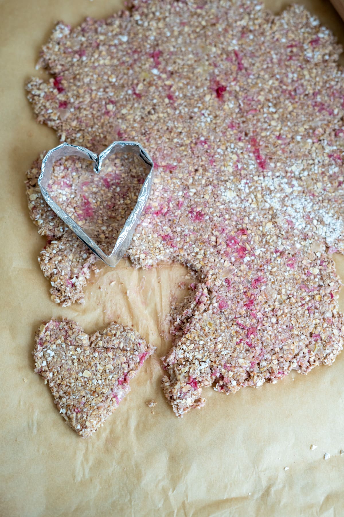 Heart-shaped biscuit cutter and cutout on rolled oat and berry dough placed on baking parchment.