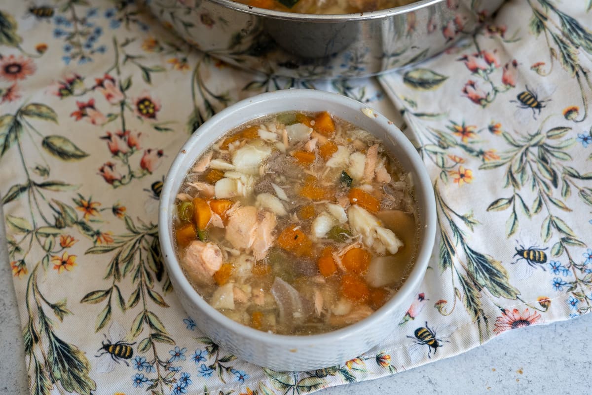 A white bowl filled with chunky soup containing pieces of fish, carrots, potatoes, and vegetables, placed on a floral-patterned cloth.