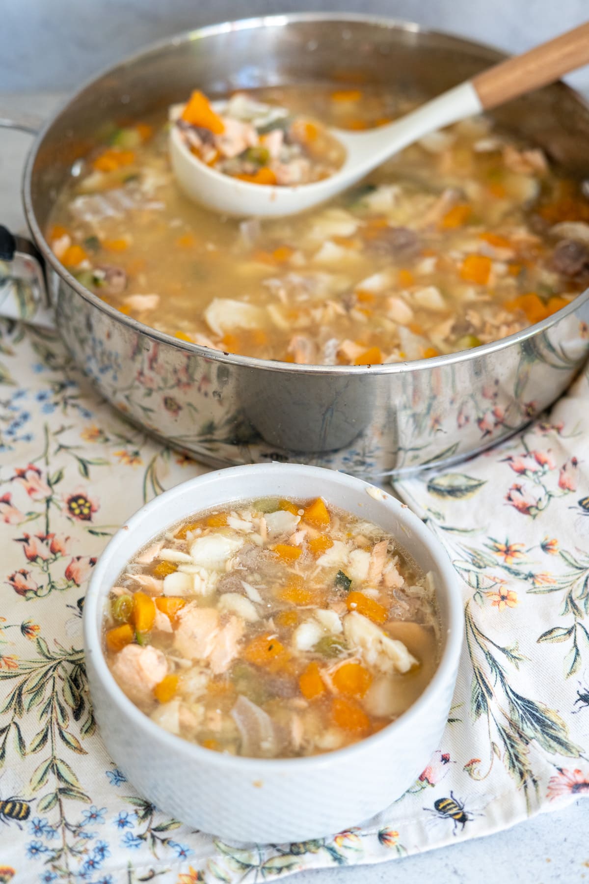 A white bowl and a ladle filled with chunky fish soup with vegetables, perfect as a wholesome fish stew for dogs, placed next to a pot of the same soup on a floral-patterned cloth.