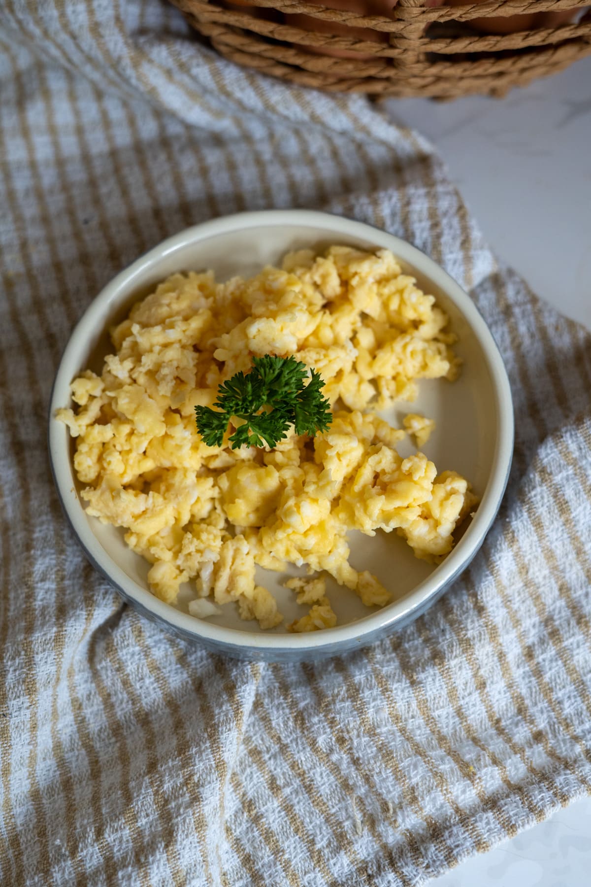 A plate of scrambled eggs for dogs, garnished with parsley, sits on a striped cloth next to a woven basket.