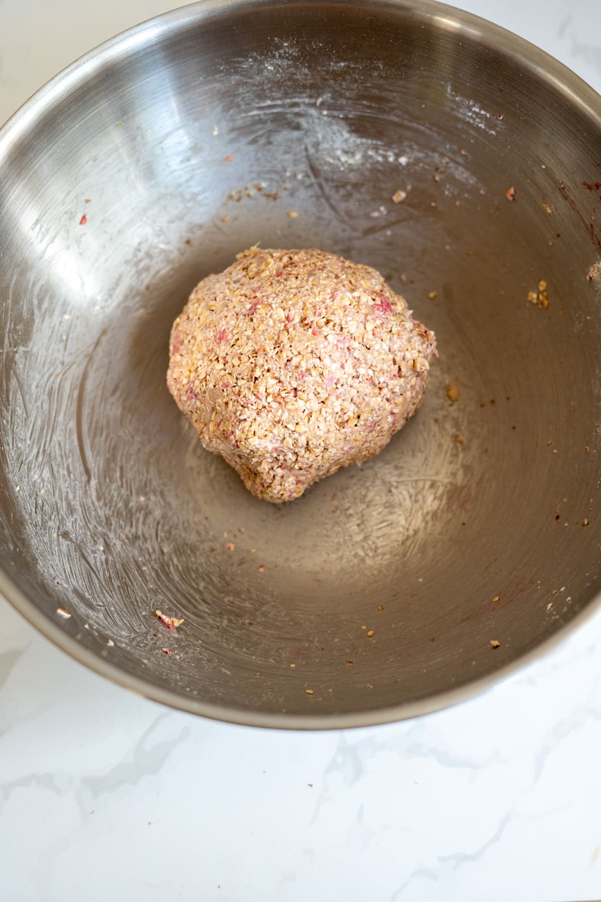 A ball of mixed ingredients sits in a large stainless steel mixing bowl on a white worktop.