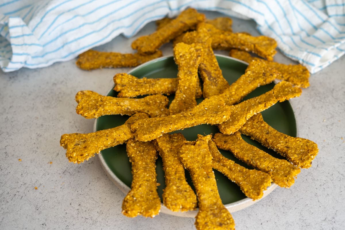 A green plate holds several bone-shaped homemade liver dog treats on a light worktop, with a striped cloth in the background.