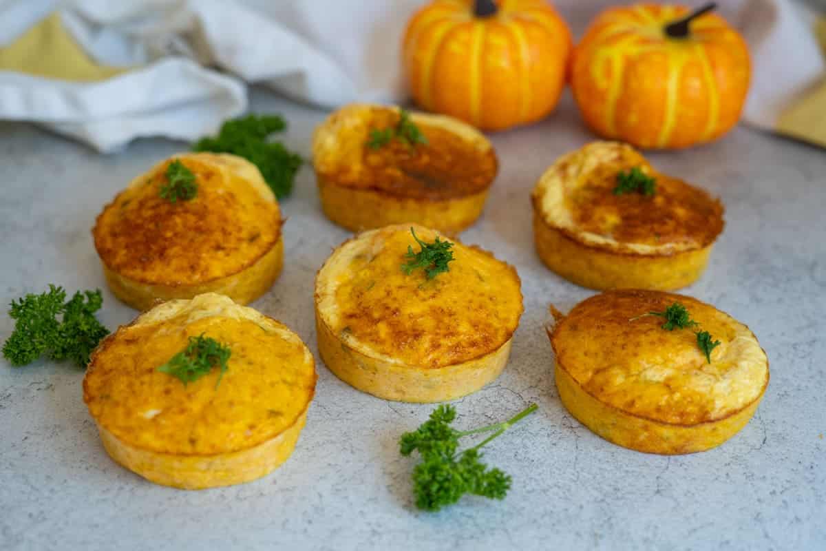 Six baked savoury pies topped with parsley are arranged on a grey surface, with two small decorative pumpkins and a white cloth in the background.