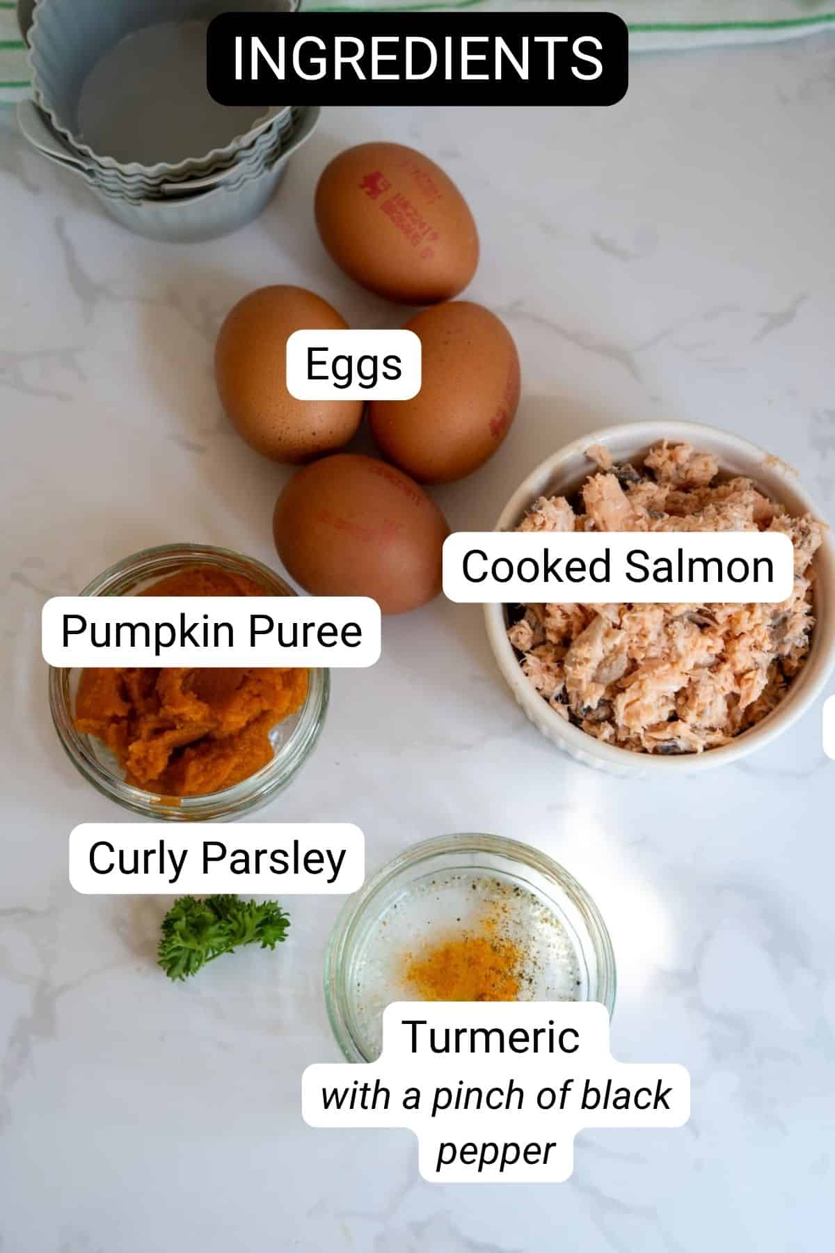 Five eggs, a bowl of cooked salmon, pumpkin purée, curly parsley, and turmeric with black pepper are arranged on a white surface. Each item is labelled.