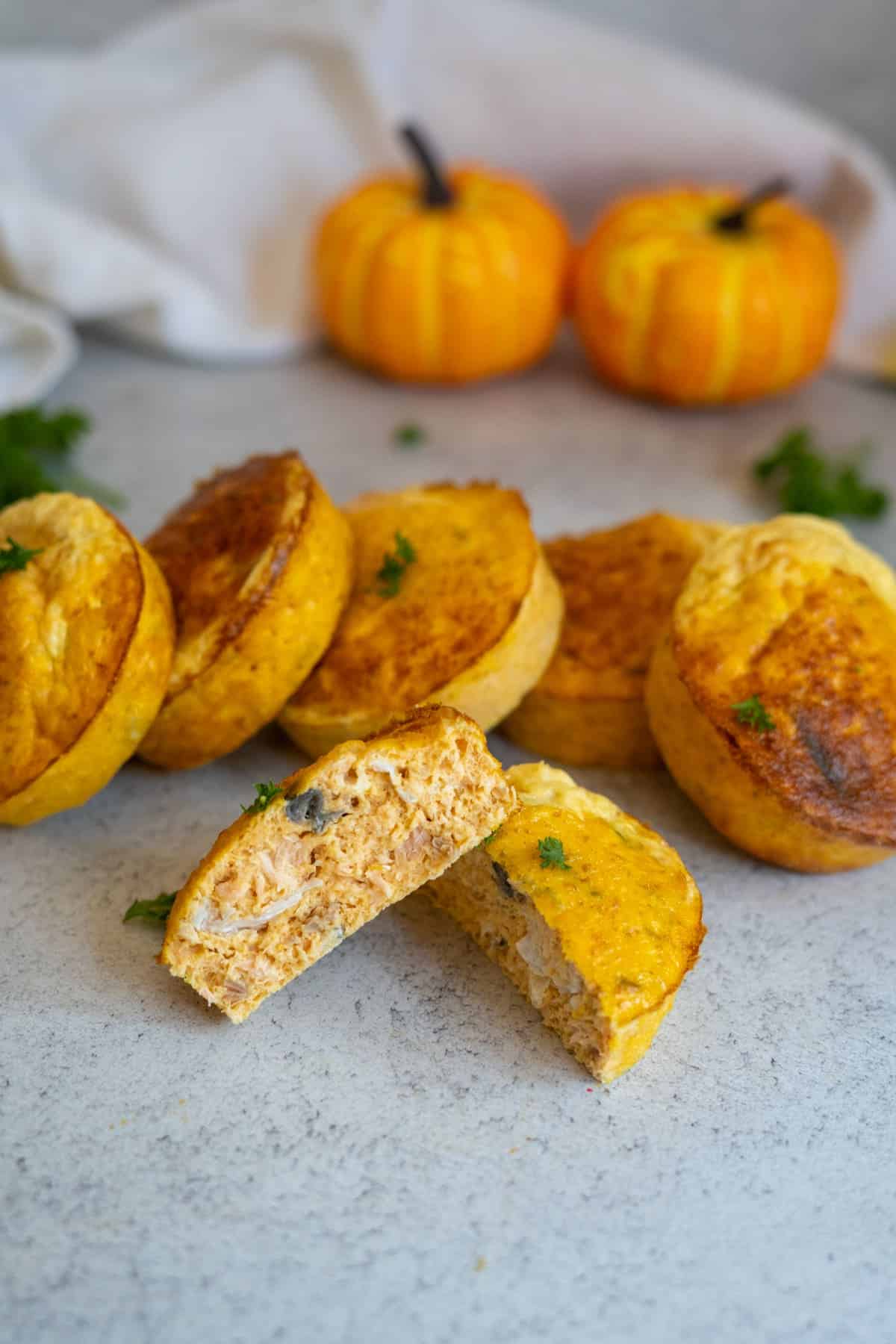 Six golden-brown, muffin-shaped salmon and pumpkin dog muffins are arranged on a light surface; one is cut in half, showing the hearty filling. Two small decorative pumpkins and parsley garnish are in the background.