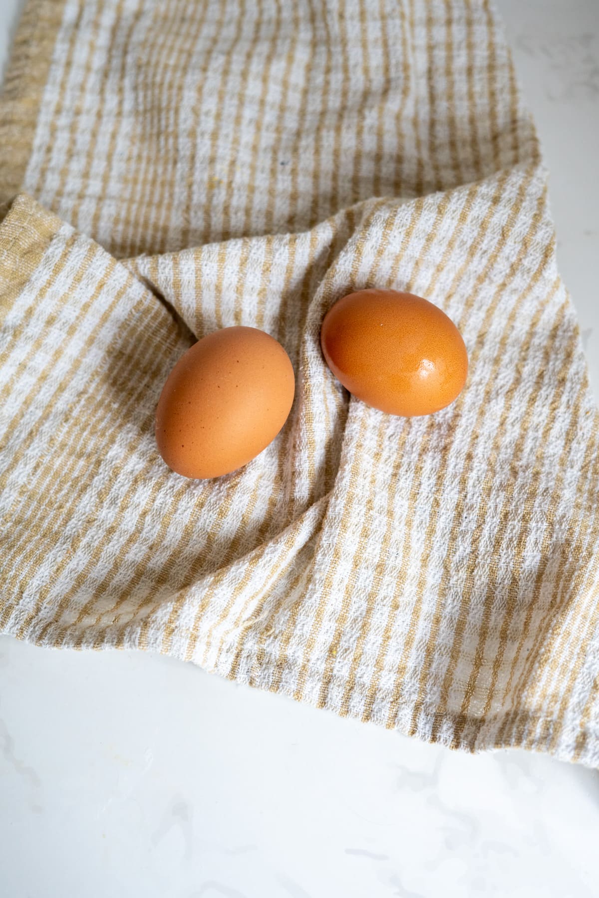 Two brown eggs rest on a beige and white striped tea towel on a light-coloured surface, ready to be whisked into scrambled eggs for dogs.