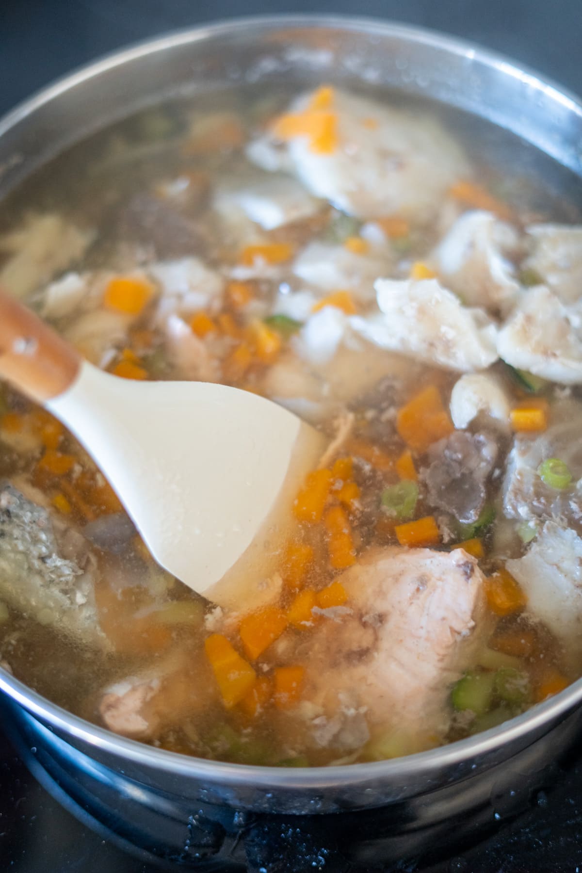A pot of soup with chunks of fish, diced carrots, and other vegetables being stirred with a white spatula—this hearty fish stew for dogs is full of wholesome ingredients your pup will love.