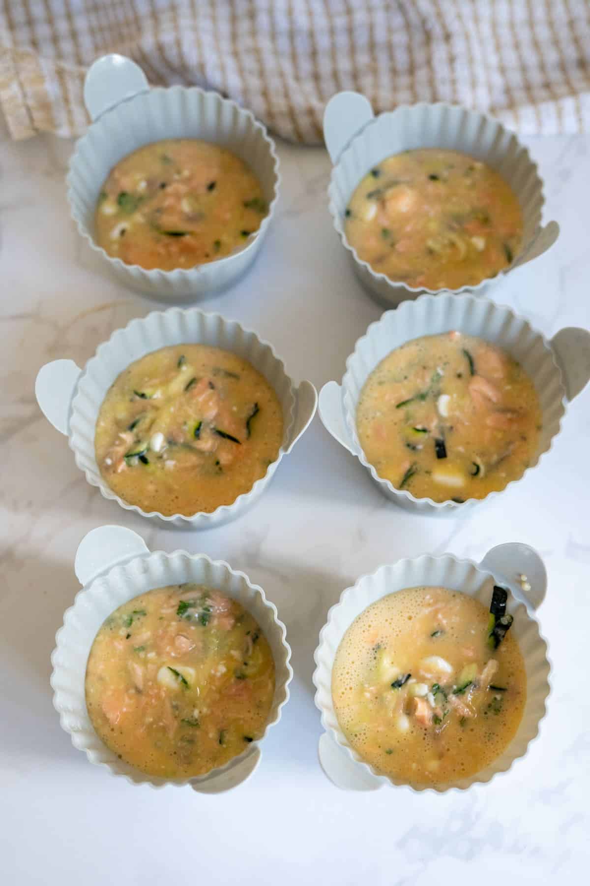 Six silicone muffin moulds filled with an uncooked egg mixture containing chopped vegetables, perfect for making Air Fryer Salmon Dog Frittata, placed on a white surface with a beige cloth in the background.