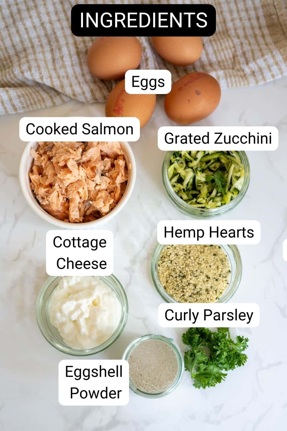 A flat lay of labelled ingredients for Air Fryer Salmon Dog Frittata, including eggs, cooked salmon, grated courgette, cottage cheese, hemp hearts, curly parsley, and eggshell powder on a light surface.