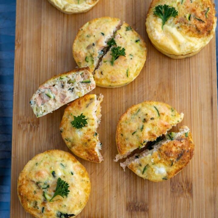 Four egg muffins with herbs are arranged on a wooden board, some cut open to reveal a filling reminiscent of an Air Fryer Salmon Dog Frittata, brimming with vegetables and meat; garnished with fresh parsley.