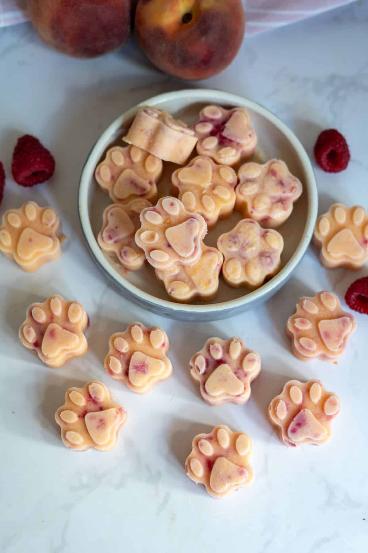 A plate and surface display Frozen Peach Raspberry Dog Treats shaped like paw prints, with peaches and raspberries nearby on a white worktop.