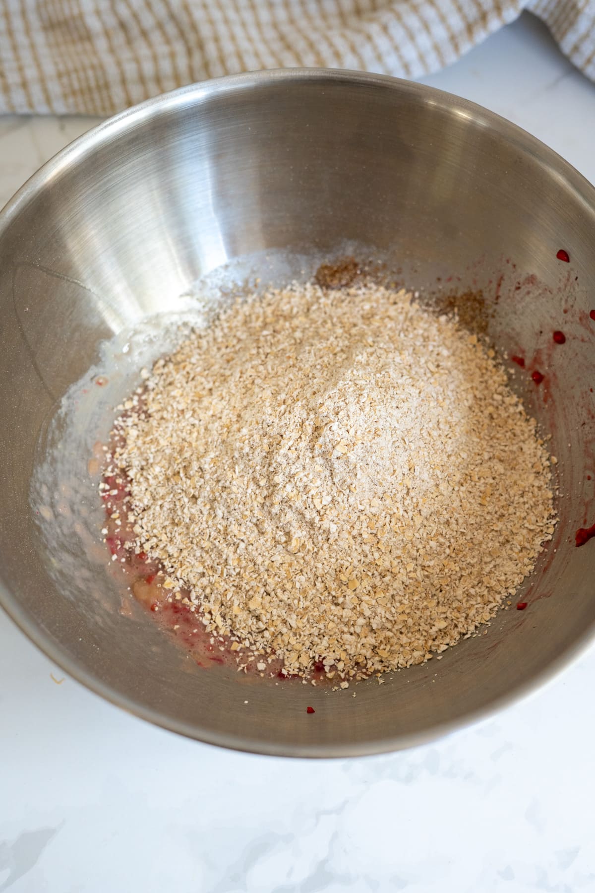 A metal mixing bowl containing oats and partially mixed ingredients, including visible red and brown flecks, sits on a white worktop.