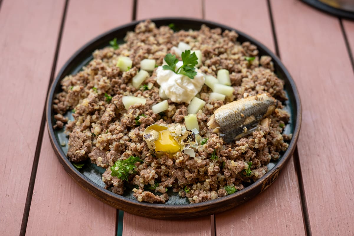 A plate of cooked minced meat mixed with quinoa and nutritious hemp hearts, topped with a sardine, diced vegetables, a dollop of soured cream, fresh parsley, and a knob of butter on a dark round plate. For pet owners wondering “can dogs eat hemp hearts,” this wholesome meal showcases how these seeds can be incorporated for added nutrition.