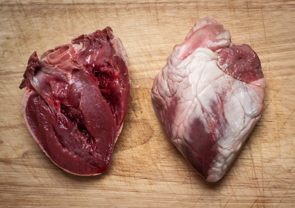 Two raw animal hearts, one whole and one cut open, displayed on a wooden chopping board.