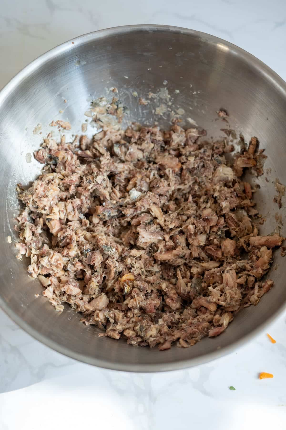 A stainless steel bowl filled with shredded cooked tuna or fish mixed with seasoning, sitting on a white worktop.
