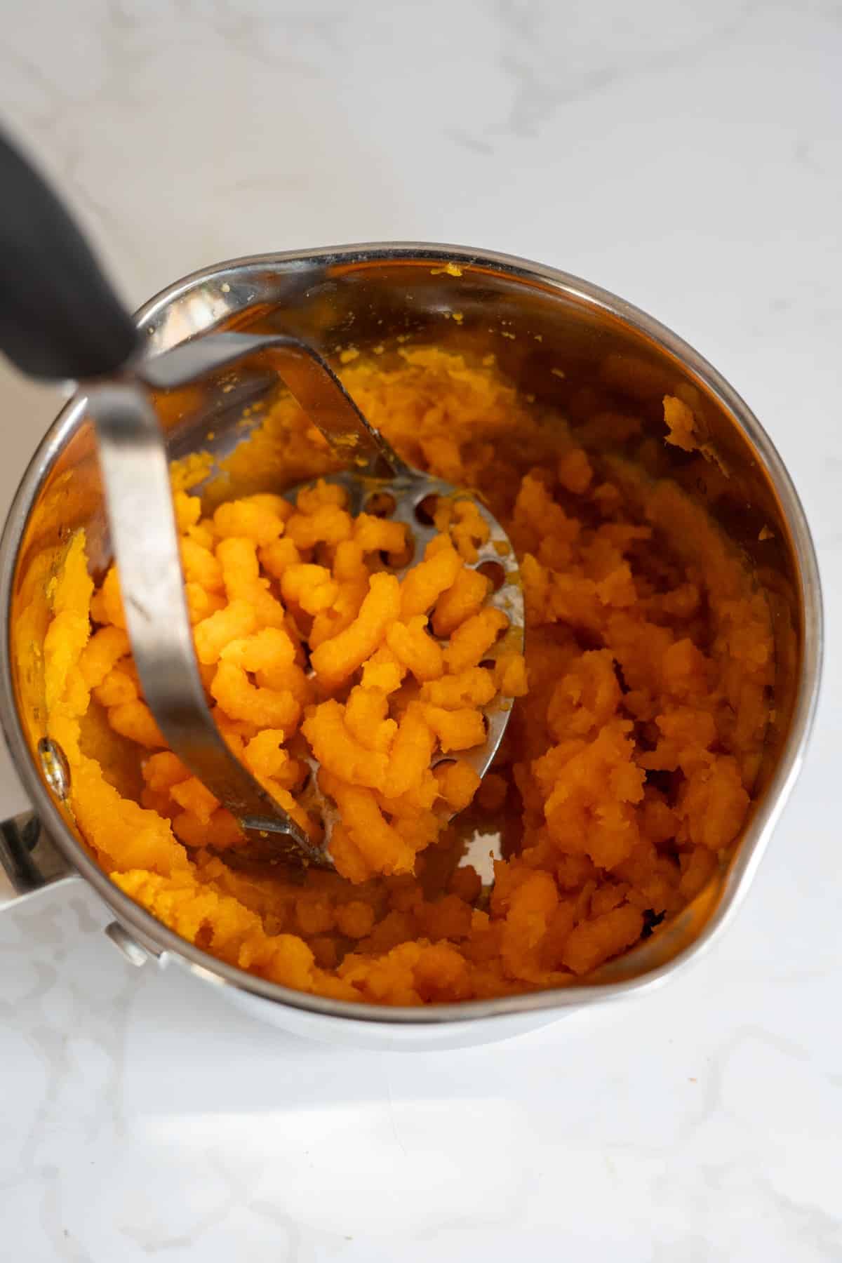 A potato masher is being used to mash cooked sweet potatoes in a stainless steel saucepan on a white surface.