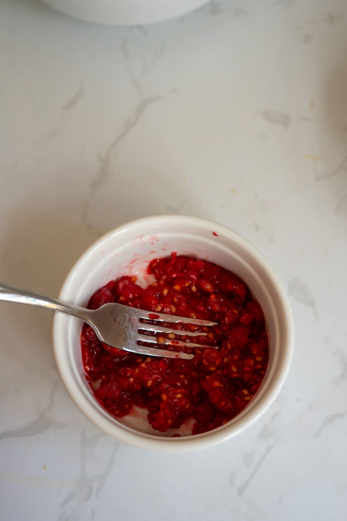 A small white bowl with mashed raspberries and a fork on a white worktop, perfect for preparing Frozen Peach Raspberry Dog Treats.