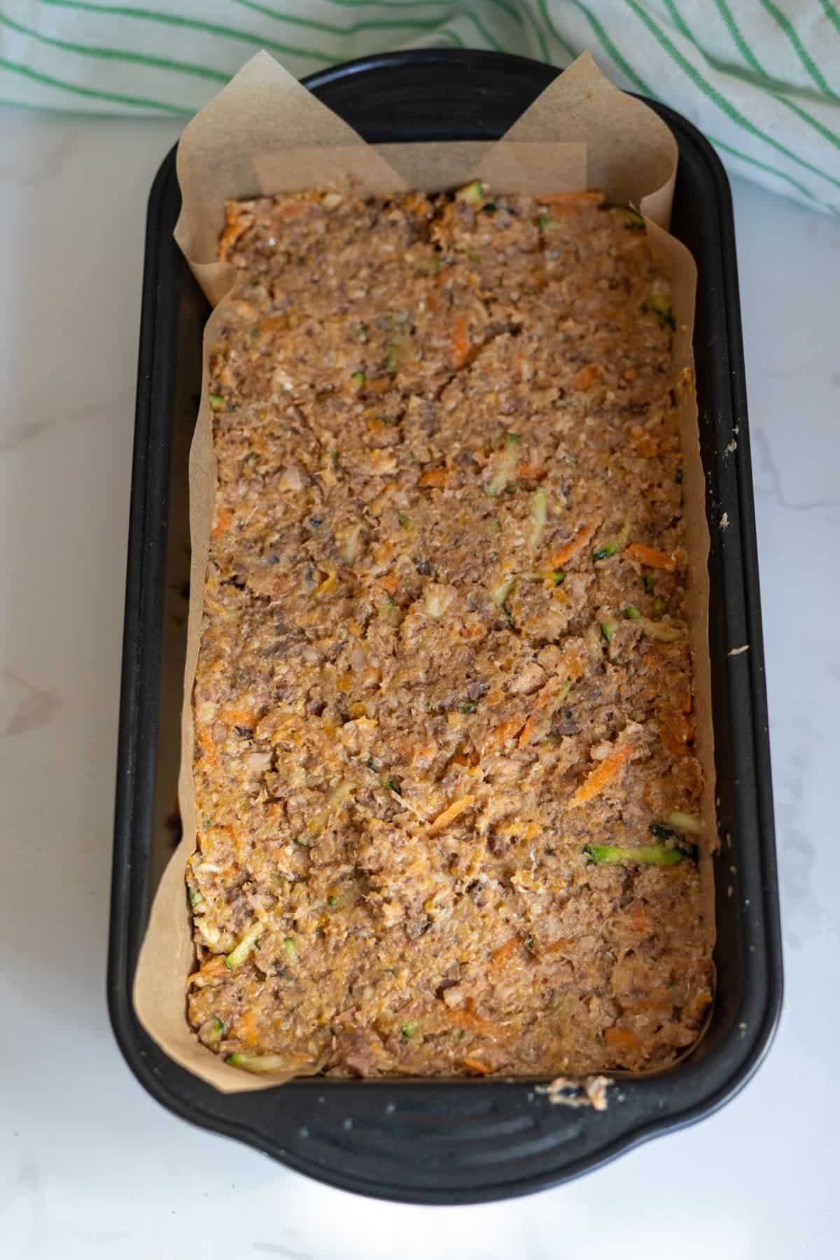 Unbaked vegetable and grain loaf mixture in a greaseproof paper-lined loaf tin, placed on a white marble surface.