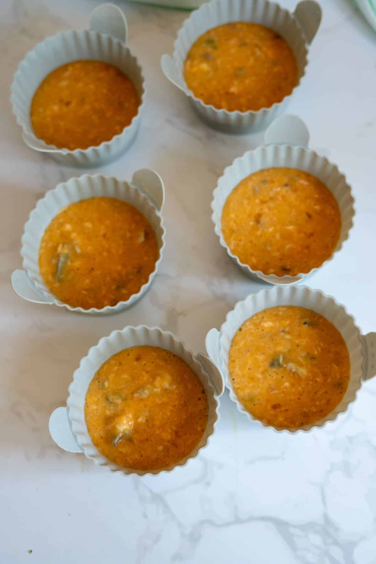 Six silicone baking cases filled with orange mixture are arranged on a white marble surface, ready for baking.