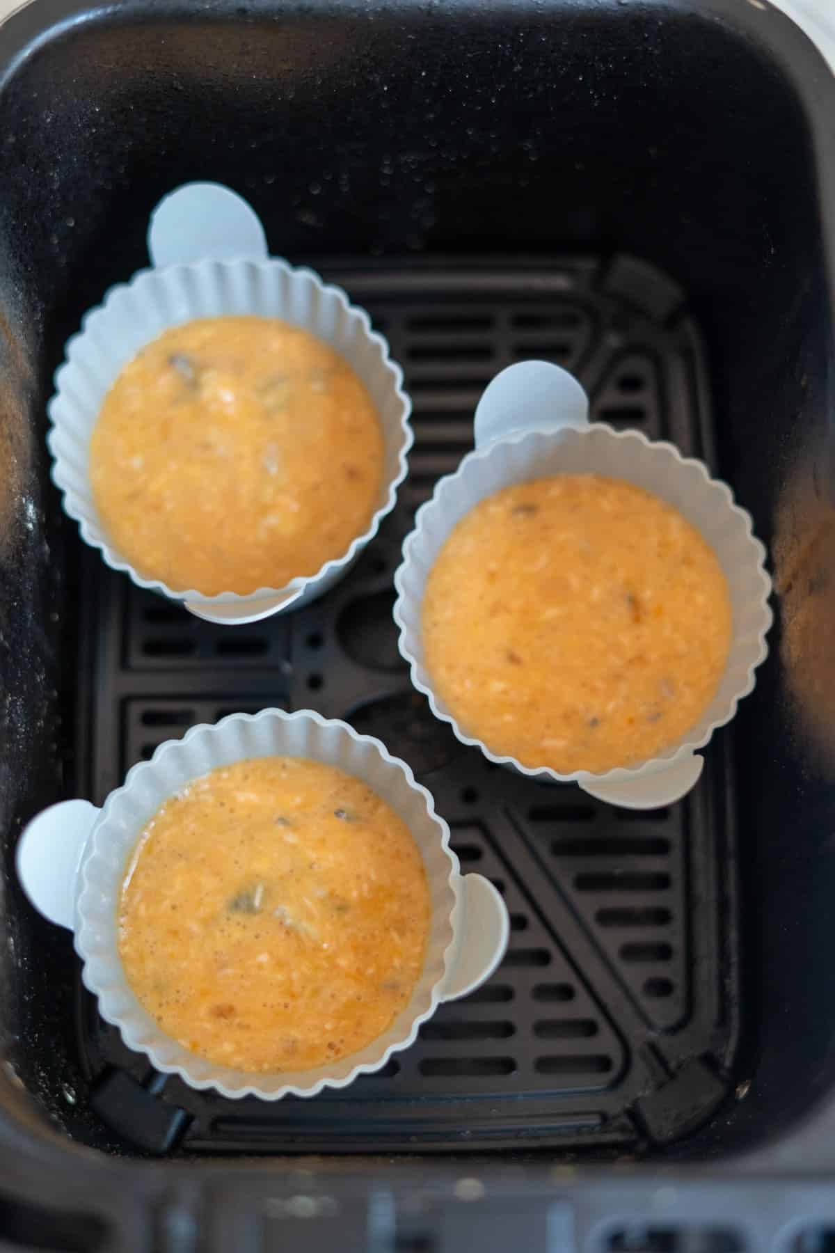 Three silicone baking cases filled with orange muffin mixture are placed inside an air fryer basket, ready for baking.