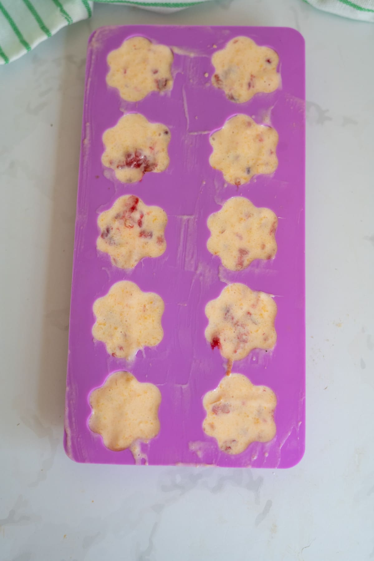A purple silicone mould with ten flower-shaped cavities is filled with a creamy mixture containing visible fruit pieces, perfect for making Frozen Peach Raspberry Dog Treats.