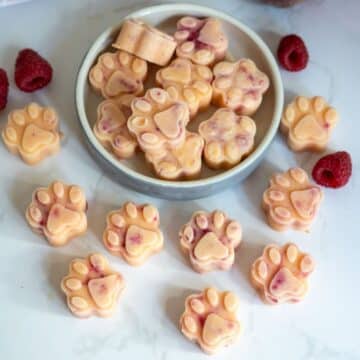 Paw-shaped iced treats with a light orange and pink colour are arranged on a plate and scattered on a white surface, with a few raspberries nearby.