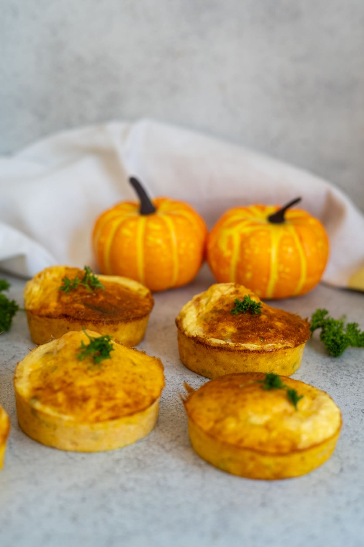 Four small savoury pies topped with parsley are arranged on a light surface, accompanied by two decorative mini pumpkins and a white cloth in the background—these salmon pumpkin dog muffins make for an inviting, festive scene.