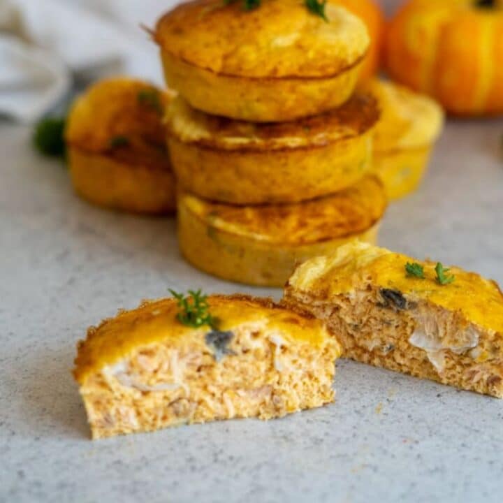 A stack of small savoury pies sits on a worktop, with one pie cut in half in front to show its filling, garnished with herbs.