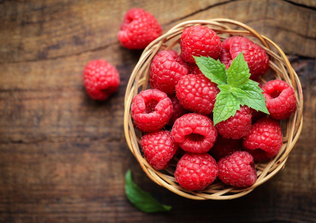A small wicker basket filled with fresh raspberries and garnished with green mint leaves sits on a wooden surface, prompting the question: can dogs eat raspberries?.