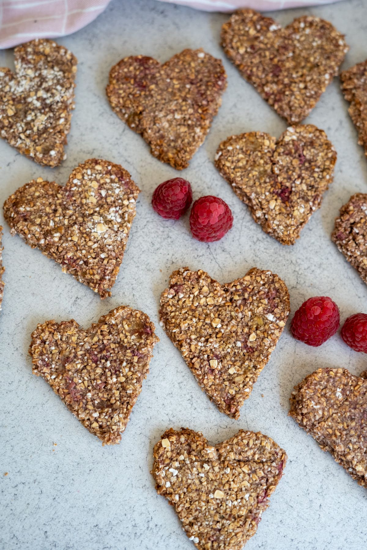 Heart-shaped raspberry oat dog treats are arranged on a light surface, with three whole raspberries placed between them.