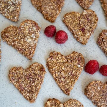 Heart-shaped oat and berry biscuits arranged on a light grey surface with three fresh raspberries placed among them.