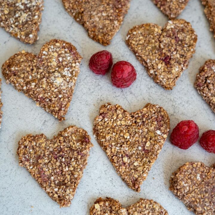Heart-shaped oat and berry biscuits arranged on a light grey surface with three fresh raspberries placed among them.