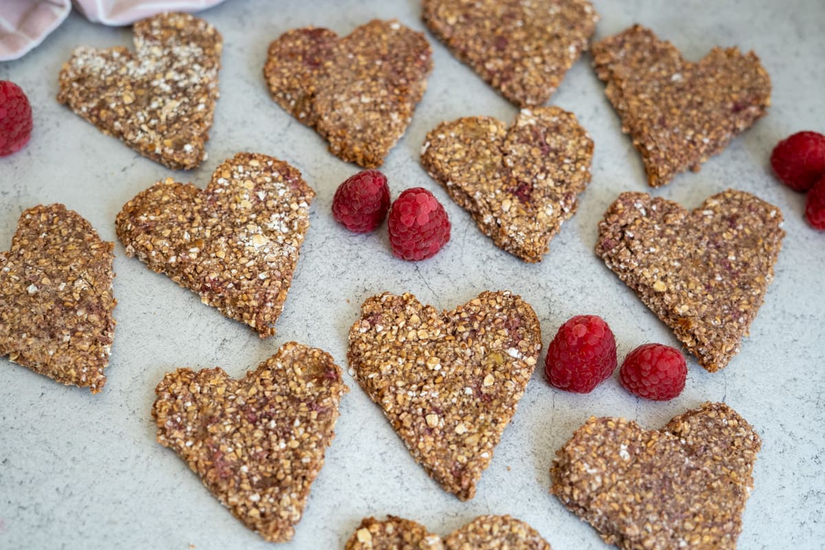 Heart-shaped oat and raspberry biscuits are arranged on a light surface, with several fresh raspberries scattered among them.