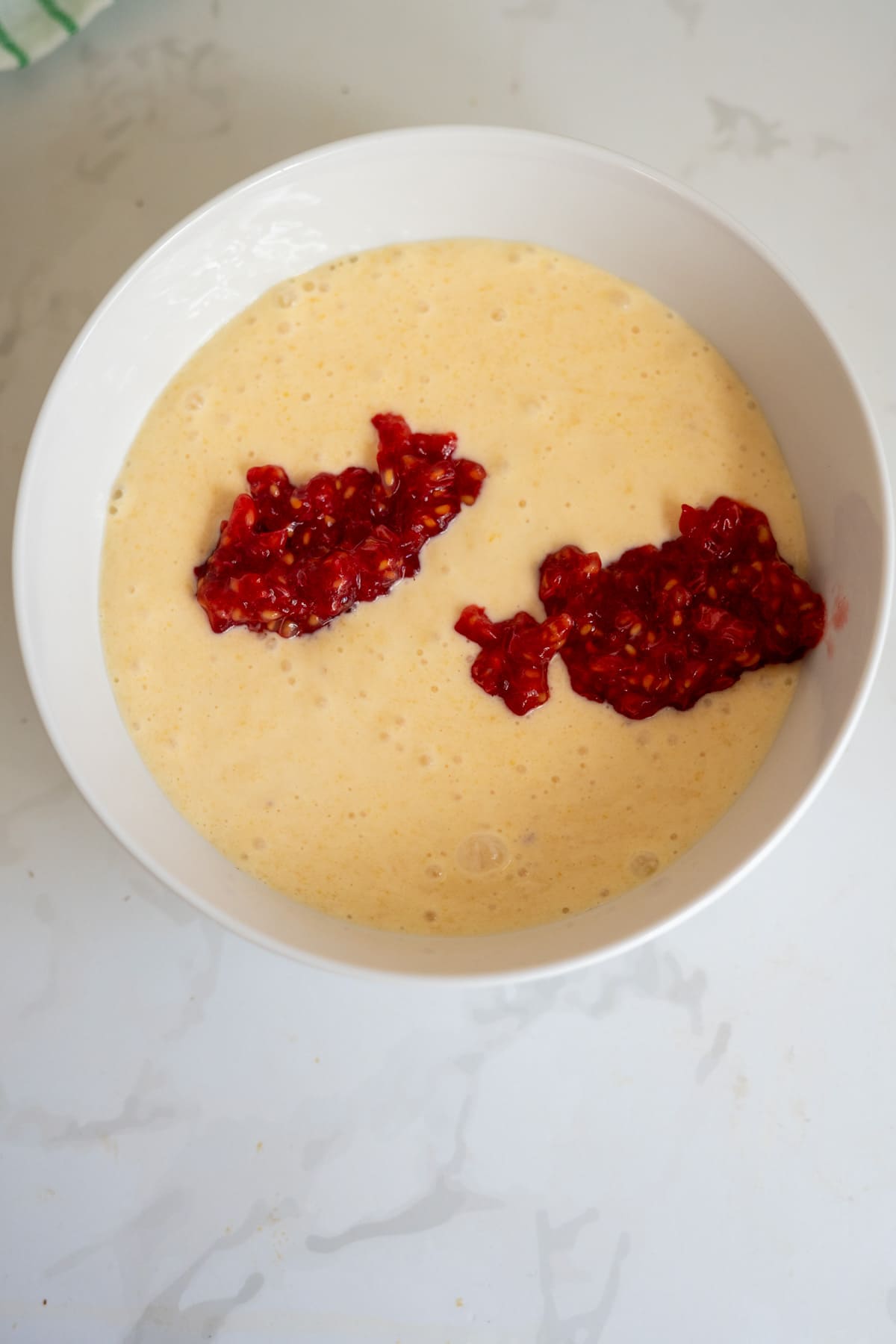 A white bowl filled with pale yellow batter, topped with two portions of chopped red fruit or berries, sits on a white marble surface—perfect for making Frozen Peach Raspberry Dog Treats.