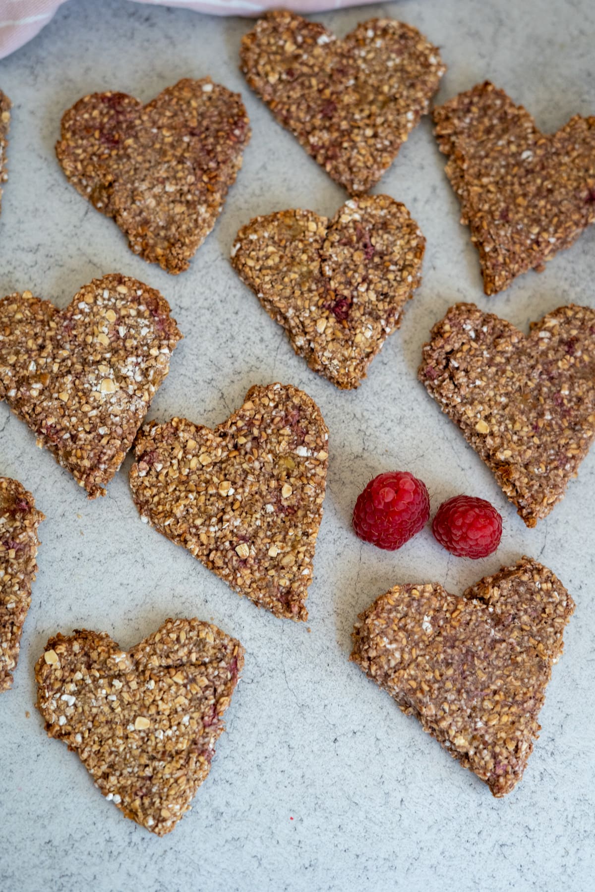 Heart-shaped oat and berry bars are arranged on a light surface next to two fresh raspberries.