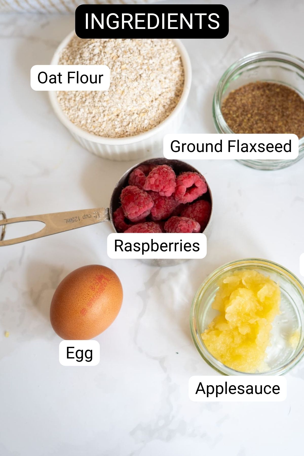 Top-down view of labelled ingredients on a worktop: oat flour, ground flaxseed, raspberries in a measuring jug, an egg, and applesauce in a small jar.