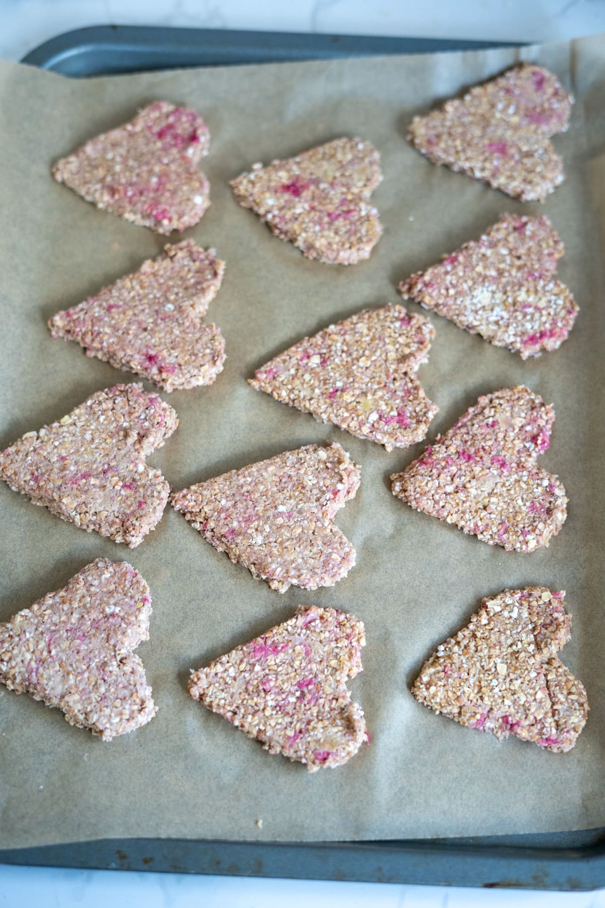 Twelve heart-shaped oat biscuits with pink specks are arranged on a baking tray lined with baking paper.