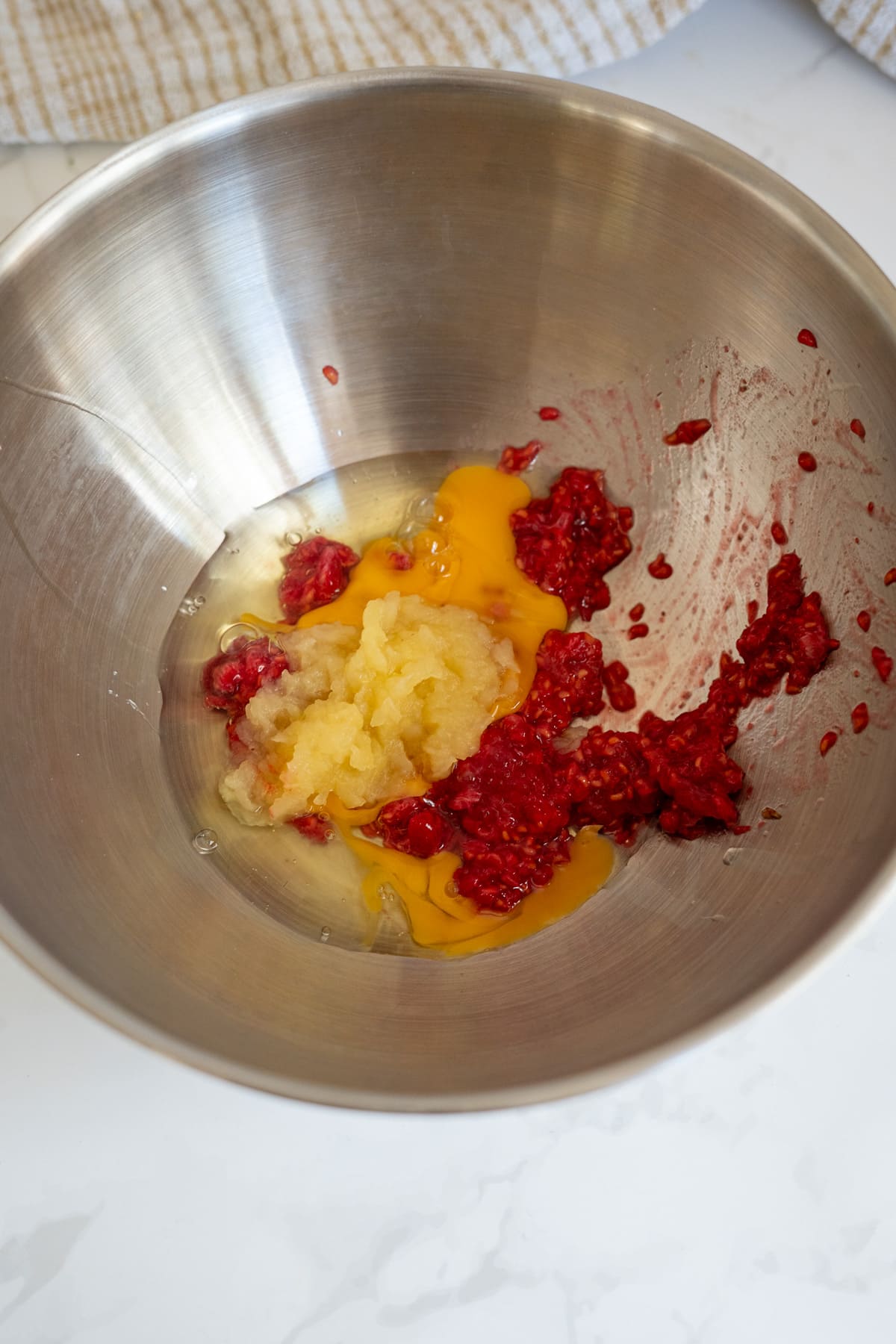 A metal mixing bowl contains cracked eggs, mashed raspberries, and what appears to be apple sauce on a white worktop.